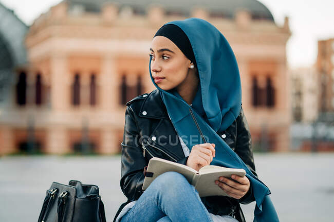 Mulher muçulmana cuidadosa em hijab escrevendo em diário enquanto sentada na rua da cidade e olhando para longe — Fotografia de Stock