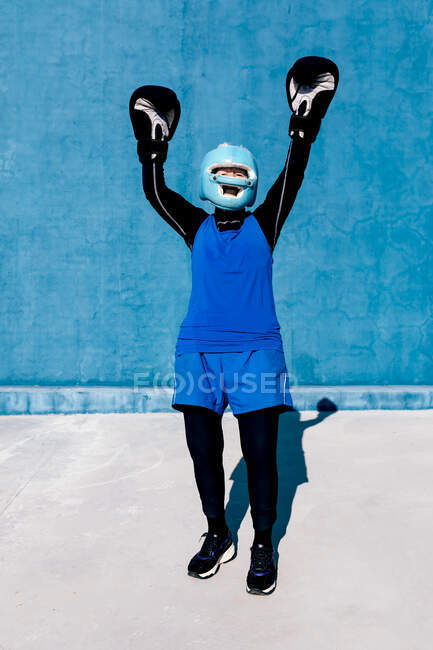 Повна довжина усміхнена зріла жінка в спортивному одязі і боксерських рукавичках, що стоять з шоломом на синій стіні з піднятими руками — стокове фото