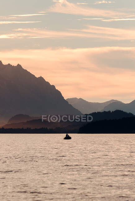 Silueta de persona anónima en moto acuática flotando en el mar tranquilo en el fondo de las montañas y el cielo puesta de sol - foto de stock