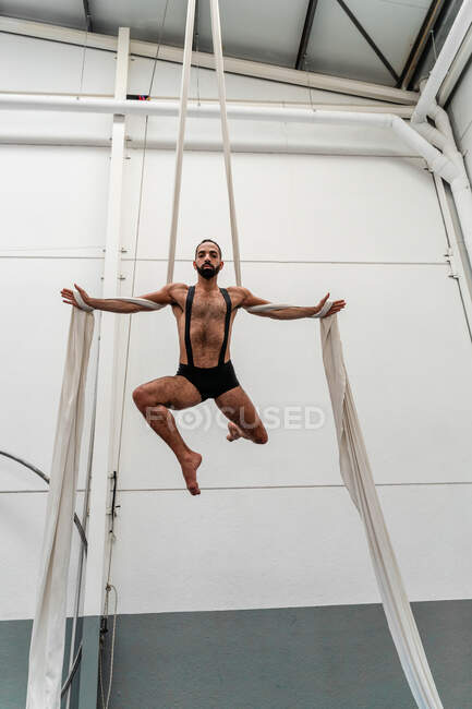Полнотелый мускулистый сильный спортсмен в шортах, выполняющий упражнения на воздушных шелках в современном световом фитнес-центре — стоковое фото