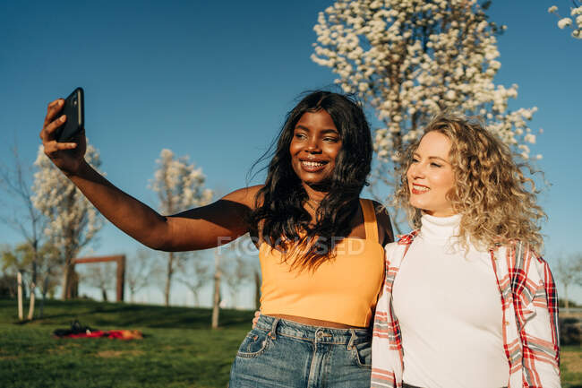 Baixo ângulo de alegre multirracial feminino melhores amigos abraçando no jardim da primavera e tomando auto-tiro no smartphone no dia ensolarado — Fotografia de Stock