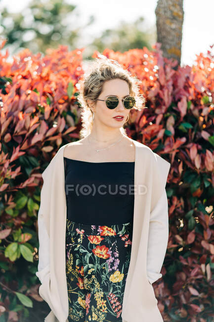 Junge Frau in trendiger Kleidung und Sonnenbrille steht an sonnigen Tagen neben roten und grünen Sträuchern im Garten — Stockfoto
