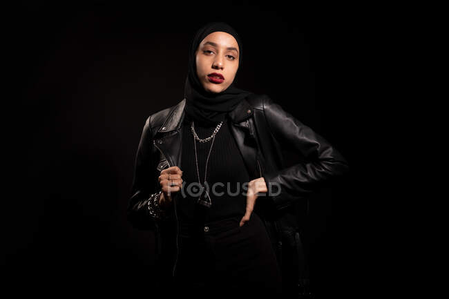 Привлекательная молодая исламская женщина в черной одежде с кожаной курткой и хиджабом, нежно смотрящая на камеру на черной студии — стоковое фото