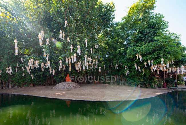 Скульптура Будды на камне под фонарями на деревьях против восточного строительства, отражающаяся в чистом пруду в Таиланде — стоковое фото