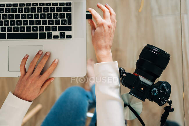 Вид зверху на врожай невідомий жінка-фотограф, що вставляє карту пам'яті фотоапарата в ноутбук, сидячи за скляним столом — стокове фото