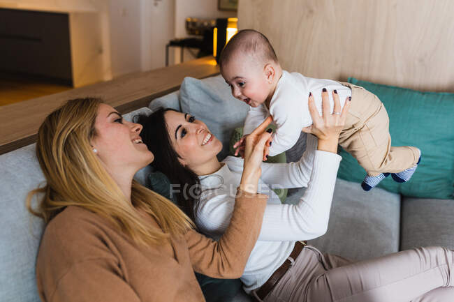 Casal alegre de mulheres homossexuais brincando com a criança bonito enquanto se divertindo e aproveitando fim de semana juntos em casa — Fotografia de Stock