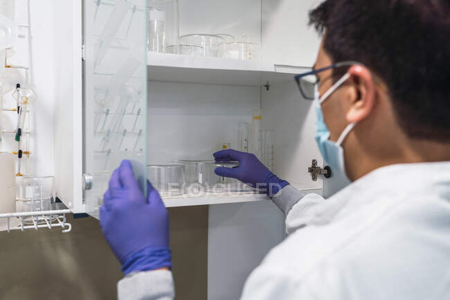 Back view crop male chemist in white robe latex gloves and face mask taking laboratory glassware from shelf while conducting research — Stock Photo