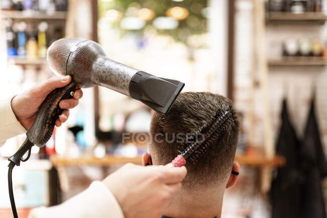 Anonymous stylist with hair dryer against man in cape in armchair in barbershop — Photo de stock