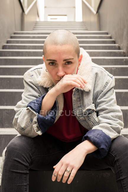 Jeune transsexuel en vêtements décontractés assis sur l'escalier entre les murs du bâtiment et regardant la caméra — Photo de stock