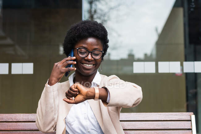 Positive schwarze Unternehmerin in formeller Kleidung spricht auf dem Handy und überprüft die Uhrzeit auf der Armbanduhr, während sie das Projekt in der Stadt diskutiert — Stockfoto