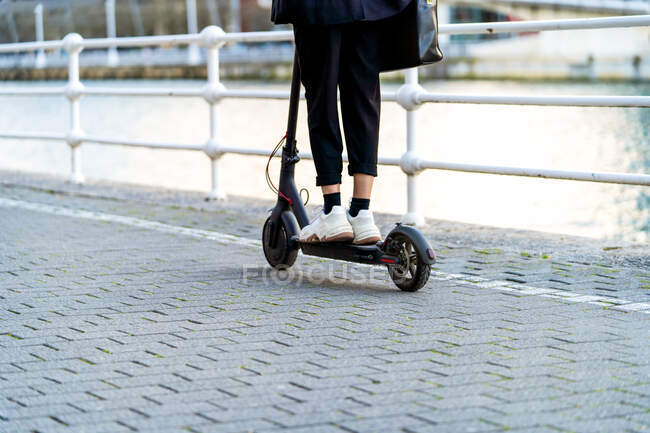 Side view of crop anonymous female in sneakers riding modern scooter on footpath against city river — Stock Photo