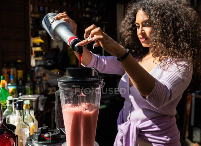 Conteúdo jovem garçom étnico feminino em uso casual adicionando xarope doce no liquidificador com smoothie de frutas enquanto mistura bebidas no bar ao ar livre ensolarado — Fotografia de Stock