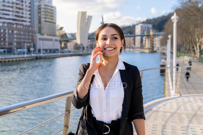 Весела молода етнічна жінка-підприємець розмовляє по мобільному телефону, дивлячись в сторону міської річки на сонячному світлі — стокове фото