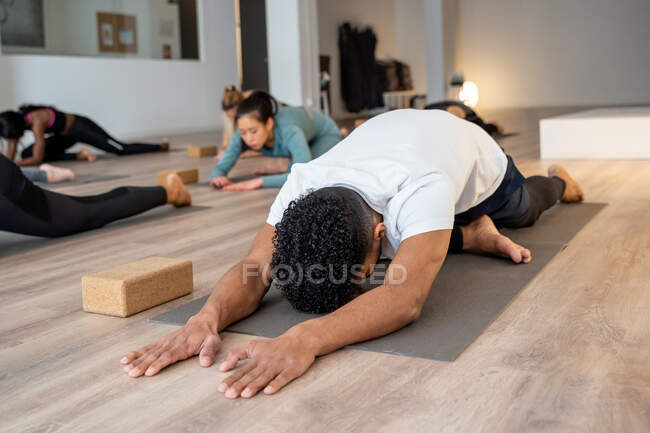 Compañía de diversas personas sentadas en esteras en Kapotasana y estirando cuerpos mientras practican yoga durante la clase en amplio estudio - foto de stock