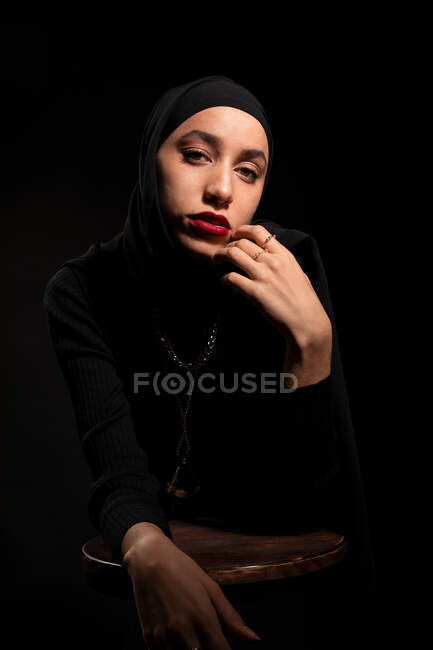 Приваблива молода ісламська жінка, одягнена в чорне вбрання і хіджаб торкається обличчя м'яко спираючись на стілець в чорній студії дивлячись на камеру — стокове фото