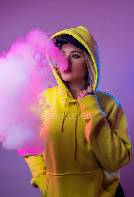 Confident female hipster in hoodie smoking e cigarette in studio on pink background and looking away — Stock Photo