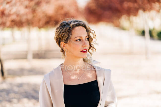 Female in elegant dress and coat walking along alley between trees and looking away on blurred background — Stock Photo