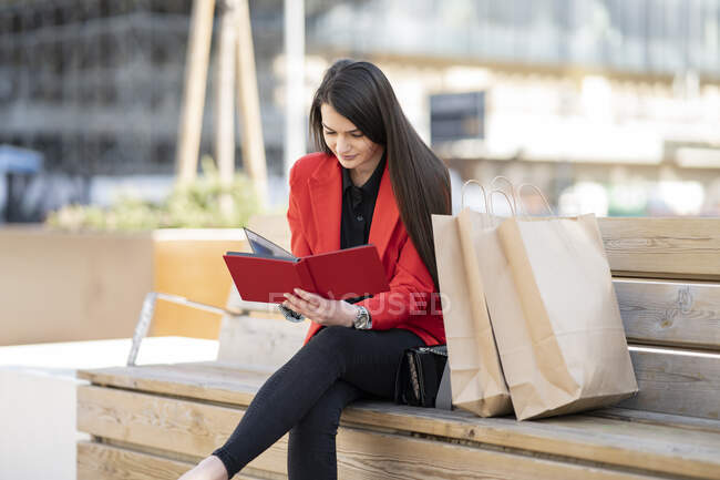 Une cliente souriante assise sur un banc avec des sacs en papier et un livre de lecture après des achats réussis en ville — Photo de stock