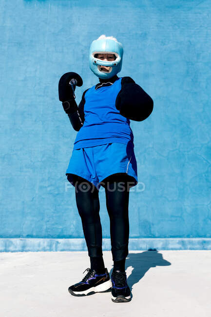 Von unten in voller Länge reife Frau in Sportbekleidung und Boxhandschuhen mit Helm vor blauer Wand stehend und in die Kamera blickend — Stockfoto