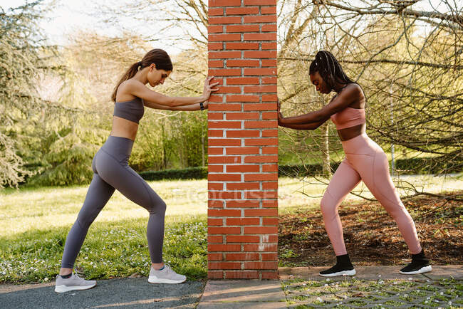 Seitenansicht junger multiethnischer Sportlerinnen in Activwear beim Training zwischen groben Pfosten, während sie im Stadtpark nach unten schauen — Stockfoto
