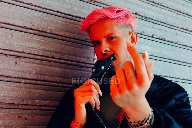 Hombre homosexual joven con tatuaje y cabello rosa en ropa de abrigo elegante mirando a la cámara contra la pared erosionada - foto de stock