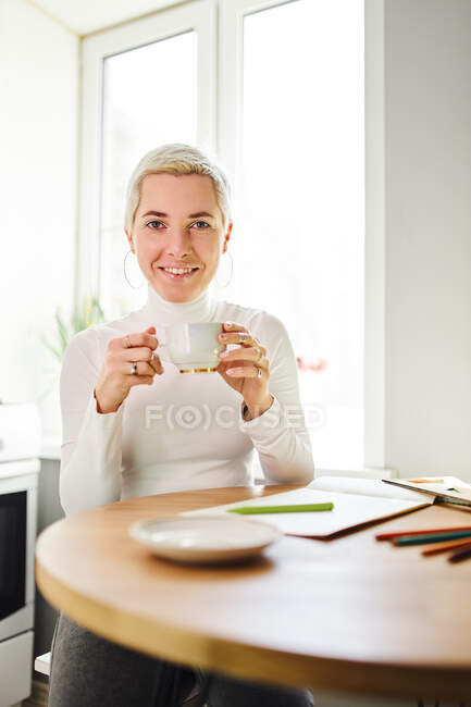 Щаслива жінка-астролог п'є гарячий напій з чашки, дивлячись на камеру вдома на сонячному світлі — стокове фото