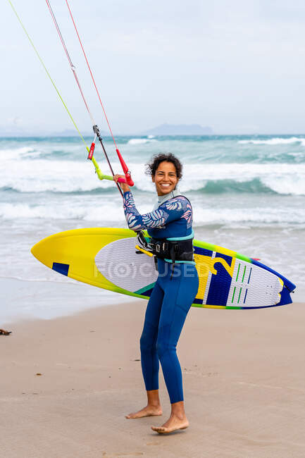 Fröhliche Kiterin im Neoprenanzug hält Kontrollstange in der Hand, während sie am sandigen Meeresufer in die Kamera blickt — Stockfoto