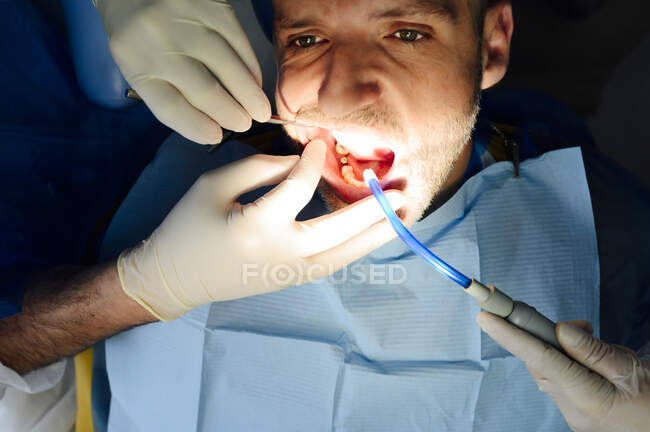 Cosecha ortodoncista anónimo en guantes de látex con compañero de trabajo comprobando los dientes de hombre maduro mirando hacia adelante en el hospital - foto de stock