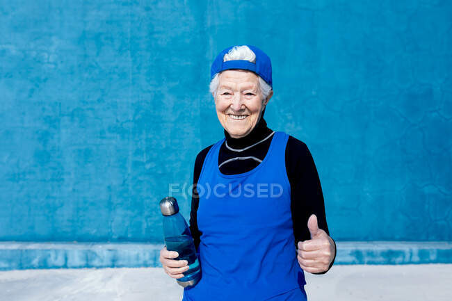 Fröhliche reife Sportlerin in Aktivkleidung und Mütze steht mit Wasserflasche in der Hand vor blauer Wand im sonnigen Outdoor-Trainingszentrum und blickt in die Kamera — Stockfoto