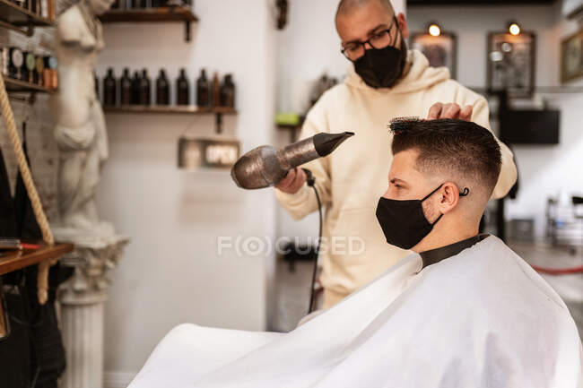 Stylist in textile mask with hair dryer against man in cape in armchair in barbershop — Fotografia de Stock