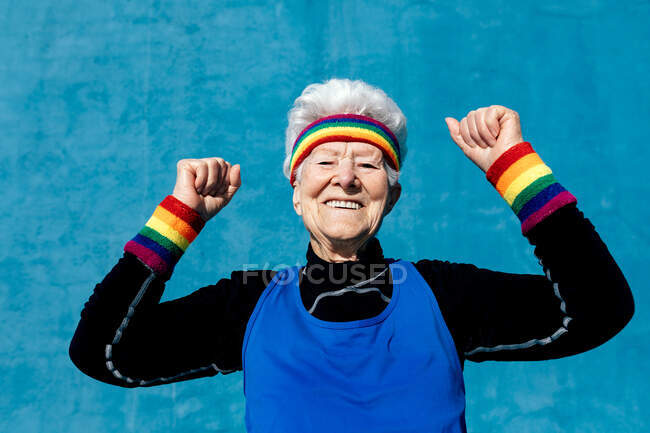 Emozionata donna anziana in abbigliamento sportivo che celebra la vittoria con pugni stretti e braccia alzate su sfondo blu in studio — Foto stock