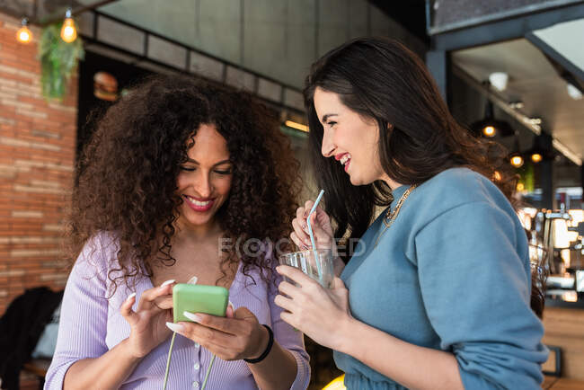 Sonrientes amigas jóvenes que usan ropa casual navegando por teléfonos móviles mientras toman un refresco en el restaurante - foto de stock