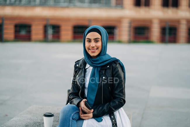 Hembra musulmana en pañuelo para la cabeza sentada en el banco con bebida para llevar mientras mira la cámara - foto de stock