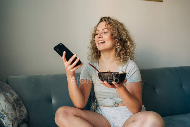 Alegre joven hembra en pantalones cortos en el teléfono móvil mientras está sentado con las piernas cruzadas en el sofá y sosteniendo tazón con sabrosa papilla - foto de stock