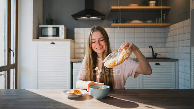 Alegre hembra vertiendo cereal crujiente en un tazón en la mesa con sabrosas galletas de avena con chips de chocolate para el desayuno en casa - foto de stock