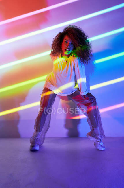 Corpo inteiro elegante confiante dançarina afro-americana com cabelo encaracolado e óculos de sol agachados olhando para a câmera em luzes de néon no estúdio de dança — Fotografia de Stock