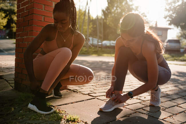 Young multiethnic female athletes tying shoelaces on footwear while squatting on urban pavement before training in back lit — Stock Photo
