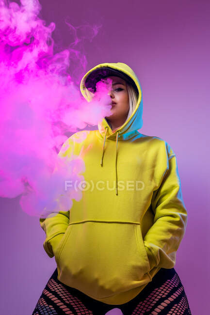 Знизу впевнена жінка-хіпстер у светрі курить електронну сигарету в студії на рожевому фоні, дивлячись на камеру — стокове фото