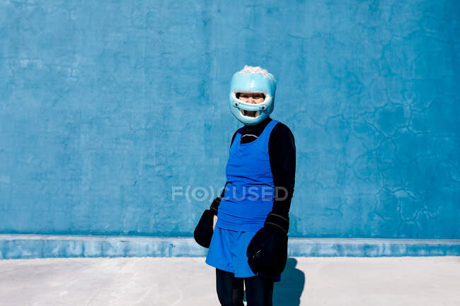 Mature female in sportswear and boxing gloves standing with helmet against blue wall and looking at camera — Stock Photo