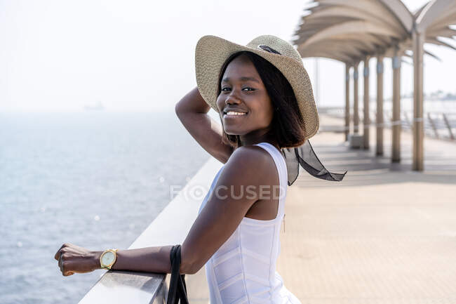 Vista lateral Afro-americano fêmea em elegante vestido branco em pé no calçadão da cidade e desfrutando de vista para o mar em tempo ensolarado — Fotografia de Stock