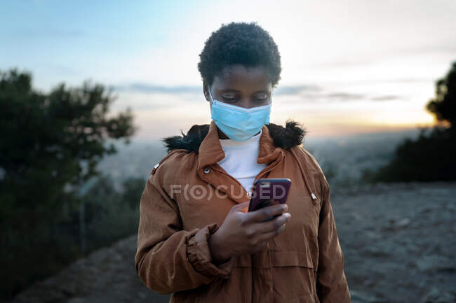Ruhige junge Afroamerikanerin in warmer Jacke und schützender Gesichtsmaske surft ihr Handy, während sie in der Dämmerung auf einem Hügel vor einer verschwommenen Stadt steht — Stockfoto