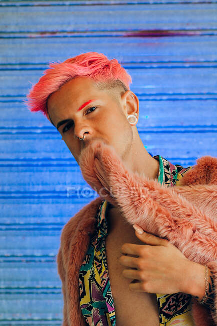 Young homosexual man in trendy wear with manicure and modern haircut biting jacket looking at camera on blue background — Stock Photo