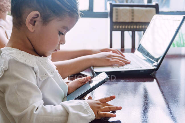 Вид збоку на етнічну маленьку дівчинку, яка переглядає мобільний телефон, сидячи за столом з матір'ю, яка набирає ноутбук вдома — стокове фото