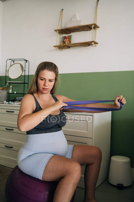 Молода майбутня жінка в спортивному одязі сидить на фітнес-кулі під час тренувань з резистентною смугою і дивиться вдома — стокове фото