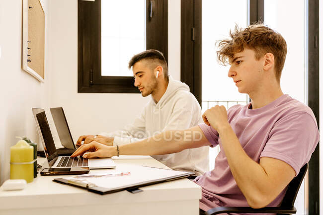 Side view of concentrated young multiethnic male colleagues in casual outfits working remotely on laptops sitting at table at home — Photo de stock