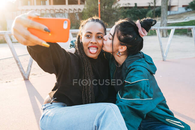 Молода гомосексуальна жінка цілує чорного коханого з язиком, знімаючи власний портрет на мобільному телефоні в місті позаду запаленого — стокове фото