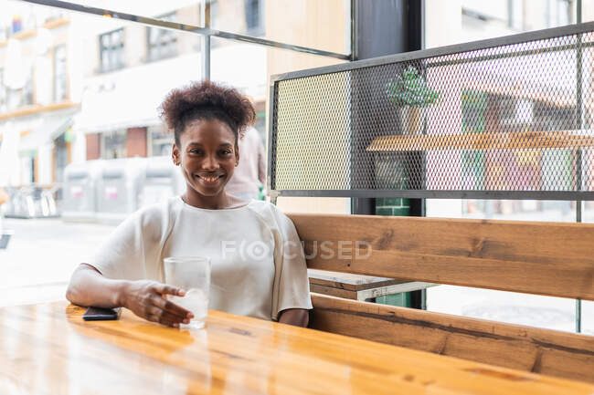 Happy young African American female in white shirt sitting with glass of icy drink in light restaurant and looking at camera with smile — Stock Photo