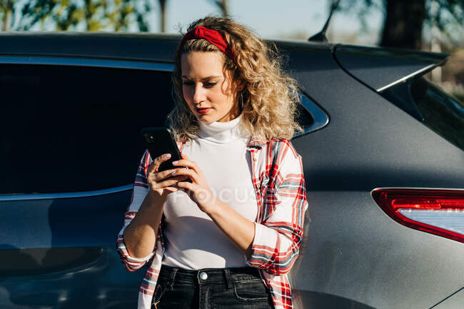 Trendy blonde female with curly hair standing near modern automobile and texting on mobile phone — Stock Photo