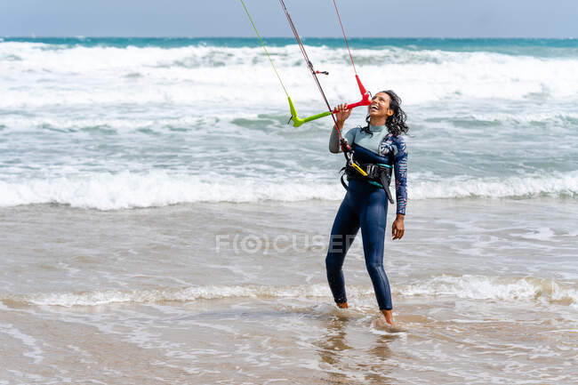 Atleta feminina em roupa de mergulho com barra de controle olhando para longe na costa arenosa contra o oceano espumoso depois de praticar kiteboarding — Fotografia de Stock