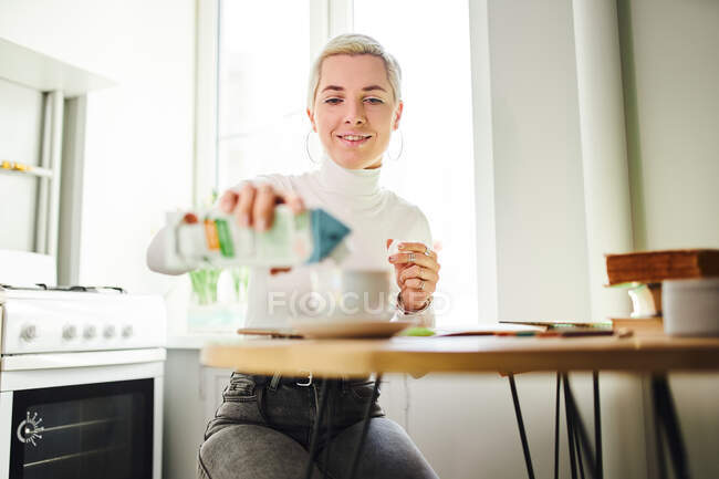 Lächelnde Astrologin gießt an sonnigen Tagen am Tisch im Haus Milch aus Karton in Heißgetränk — Stockfoto